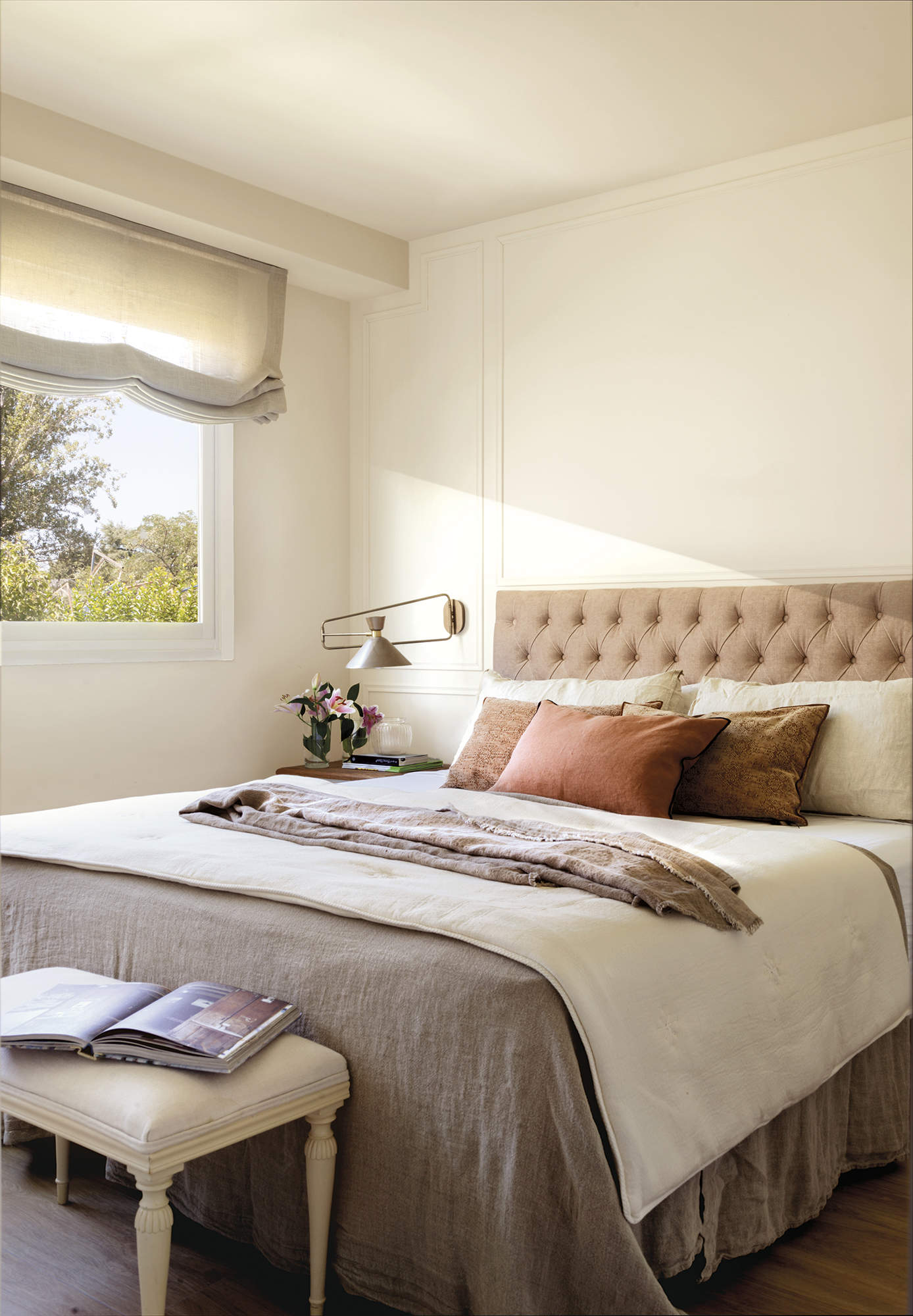 Dormitorio principal con cama con cabecero tapizado y aplique, estor gris, pared con molduras, banco estilo francés tapizado, ropa de cama gris, flores, suelo de madera